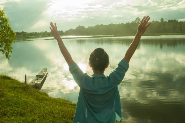 Woman standing in front of a lake with her arms raised outwards above her head. The lake's calm water is reflecting the sun and clouds.