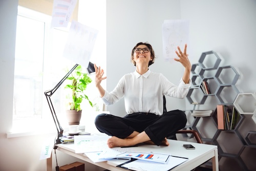 Authorpreneur's Manifesto Young beautiful business woman with glasses smiling and throwing up papers sitting cross-legged on a desk in her office
