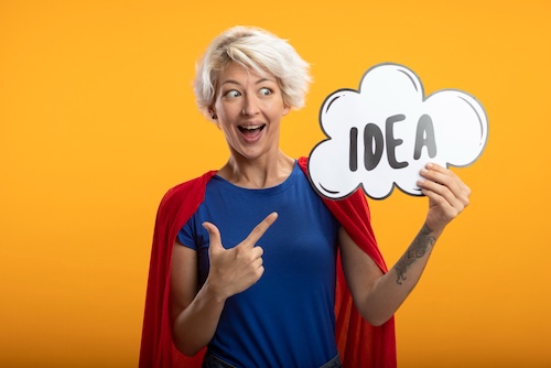 Excited superwoman with red cape holds and points at idea bubble isolated on orange wall