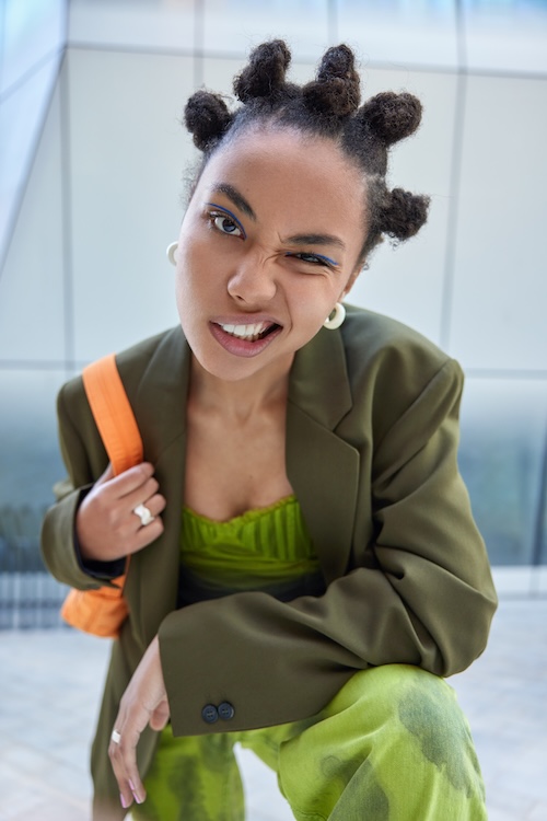 Trendy hipster girl winks eye makes cheeky expression dressed in green fashionable costume carries bag has bun hairstyle vivid makeup poses outdoors against blurred background. Urban lifestyle