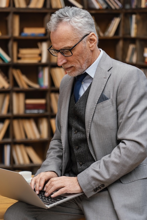 Older man writing book with AI on a laptop in a library.