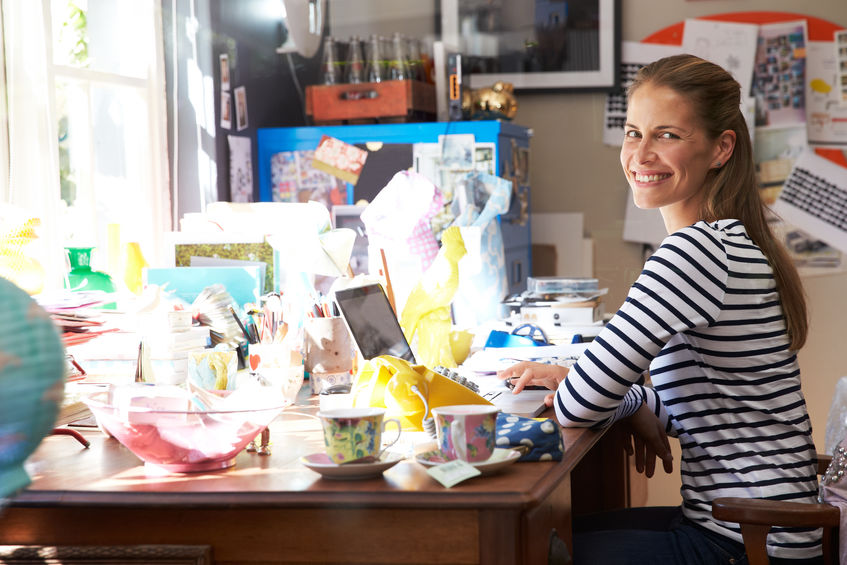 Woman running small business from her home office. She is smiling at her desk with a lot of clutter and objects.