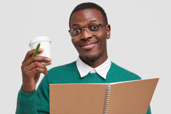 Young man with stubble, wears square spectacles, holds takeaway coffee, pen and notebook, pleased with good working result, isolated over white background.