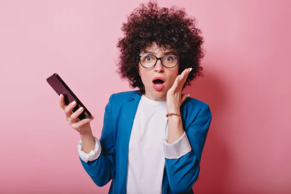 Surprised woman with wavy short hair dressed blue jacket holds smartphone with open mouth on the pink background.